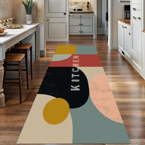 May include: A rectangular kitchen rug with a geometric design. The rug features a color block pattern with mustard yellow, red, black, and teal shapes. The word "KITCHEN" is printed vertically in white. The rug is placed on a wooden floor.