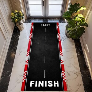May include: Black rectangular doormat with a race track design. The mat features a black surface with white dashed lines, red and white chequered borders, and the words "START" and "FINISH" in white.