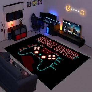 Może przedstawiać: Czarny prostokątny dywan z dużą, stylizowaną grafiką kontrolera do gier w kolorze czarnym, czerwonym i turkusowym. Nad kontrolerem widnieje napis "GAME OVER". Dywan znajduje się w pokoju gier z biurkiem, telewizorem i sofą.