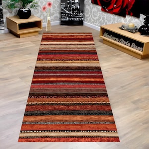 Puede incluir: Una alfombra rectangular con rayas horizontales en tonos rojos, naranjas, marrones y beige. La alfombra tiene un aspecto texturizado con patrones variados dentro de las rayas. Se coloca sobre un suelo de madera clara. Un elemento de decoración del hogar.