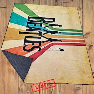 May include: A rectangular rug featuring the word "BEATLES" in large black letters, with a graphic of the band members walking. The background has colorful stripes in shades of green, blue, orange, and red. The rug has a non-slip backing.