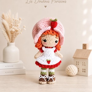 May include: A crocheted doll with orange hair wearing a pink strawberry-themed hat, a white apron with a small strawberry motif, a red skirt, green and white striped socks, and brown shoes. The text Les Doudous Parisiens is visible above.