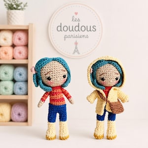 Peut inclure: Deux poupées crochetées à la main avec des cheveux bleus, l'une portant une chemise rayée rouge et orange et un pantalon bleu, l'autre un manteau jaune. Les poupées ont la peau bronzée et se tiennent devant une enseigne "les doudous parisiens".