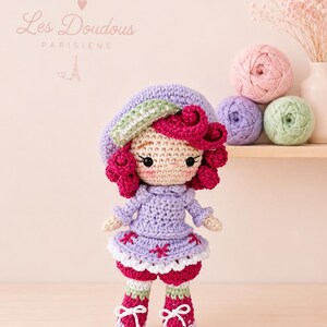 Peut inclure: Une poupée crochetée aux cheveux bouclés magenta, portant un béret lavande avec une bordure verte, un haut lavande à manches longues, une jupe lavande avec des broderies florales roses et des bottines magenta à lacets blancs. Texte Les Doudous Parisiens.