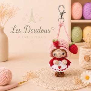 Peut inclure: Un porte-clés fait main représentant une petite fille fraise au crochet. La poupée porte une robe rouge et blanche et un chapeau rose avec une fraise. Le porte-clés a un fermoir argenté et un cordon rose. Le texte "Les Doudous Parisiens" est visible.