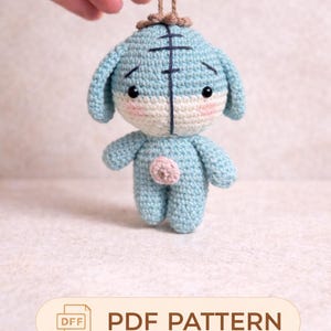 Peut inclure: Un animal en crochet bleu clair fait à la main, avec un ventre rose clair et une ficelle marron pour l'accrocher. Le visage est moitié bleu clair et moitié blanc. Le texte "PDF PATTERN" est affiché sur une bannière beige.