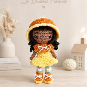 Peut inclure: Poupée au crochet faite à la main, avec une peau foncée, des cheveux noirs et une robe jaune. La poupée porte un chapeau et des chaussures orange, avec des accents blancs. Le texte "Les Doudous Parisiens" est au-dessus de la poupée.