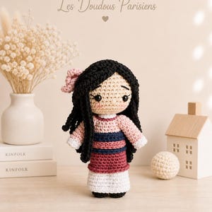 Peut inclure: Poupée au crochet faite à la main, cheveux noirs, nœud rose, robe rose, bleue et bordeaux. La poupée est posée sur une surface en bois. Le texte "Les Doudous Parisiens" est au-dessus.