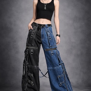 May include: A black tank top paired with two-tone denim cargo pants. The pants feature a black leg and a blue leg, with multiple pockets, zippers, and metal hardware. The model is wearing black platform boots. The background is a solid gray.