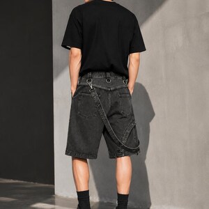 Op de afbeelding: Een persoon draagt een zwart t-shirt, een zwarte spijkerbroek met breteldetails, zwarte sokken en zwarte plateausneakers. De shorts hebben metalen ringaccenten en een uniek bandontwerp. De outfit is helemaal zwart.