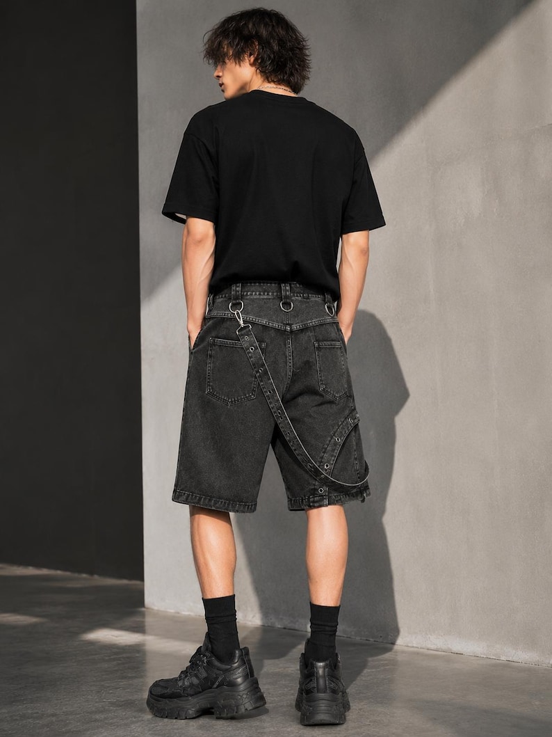 Op de afbeelding: Een persoon draagt een zwart t-shirt, een zwarte spijkerbroek met breteldetails, zwarte sokken en zwarte plateausneakers. De shorts hebben metalen ringaccenten en een uniek bandontwerp. De outfit is helemaal zwart.