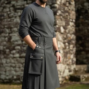 Op de afbeelding: Een antracietgrijze kilt met een geplooid ontwerp en een zak aan de voorkant. De kilt heeft een riem met metalen hardware. De persoon draagt een grijze top met lange mouwen en een horloge.