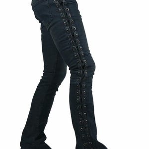 May include: Dark wash denim jeans featuring black lacing detail along the outer seams. The jeans have a slight flare at the bottom and are worn with black boots. The style is alternative and edgy, with a focus on the lacing.