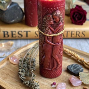 Puede incluir: Una vela de pilar roja con un dise&ntilde;o tallado de dos figuras abraz&aacute;ndose, envuelta con hilo dorado. La vela est&aacute; encendida, con una llama visible. La vela est&aacute; sobre una bandeja de madera con hierbas secas y cristales.