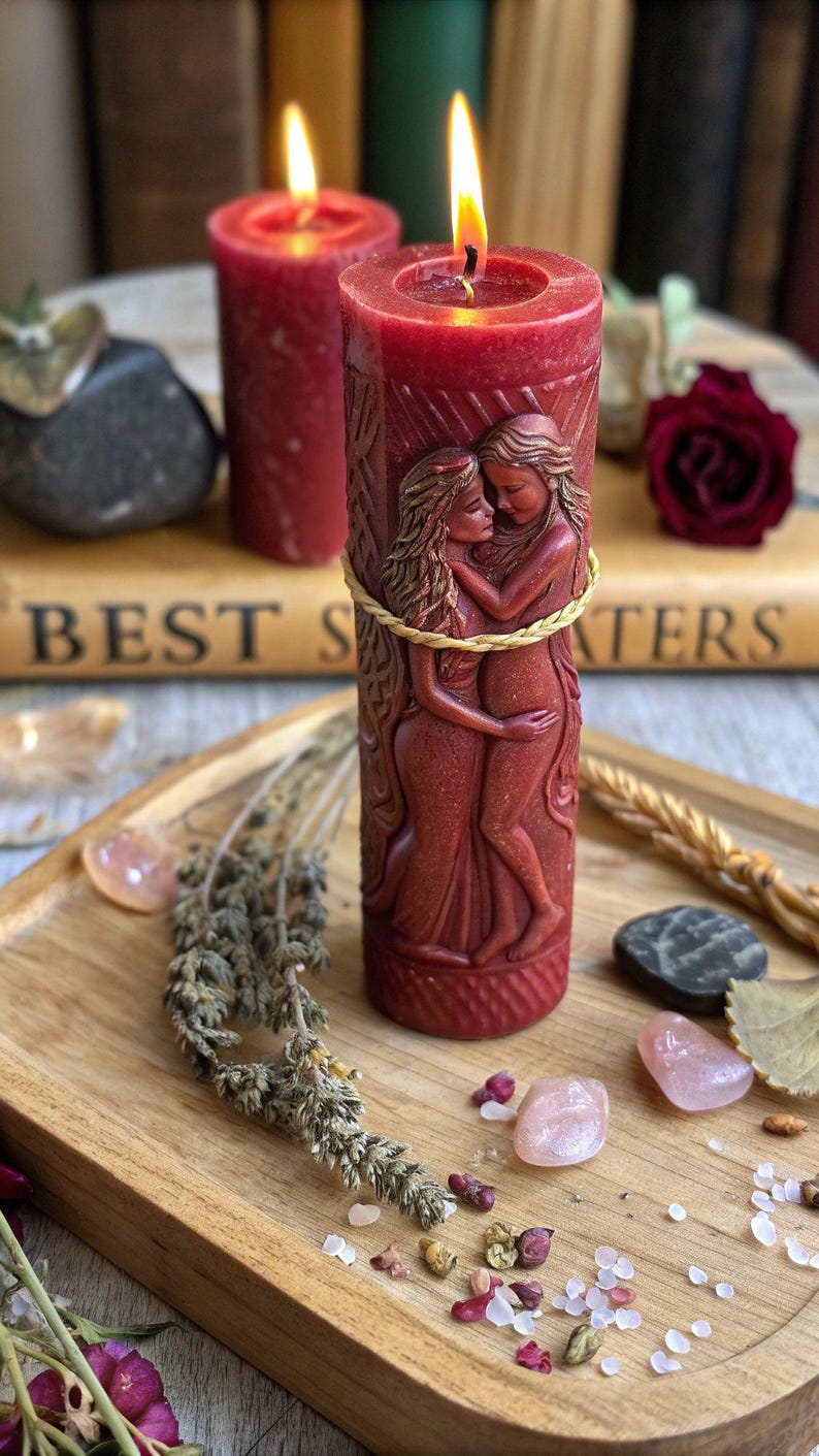Puede incluir: Una vela de pilar roja con un dise&ntilde;o tallado de dos figuras abraz&aacute;ndose, envuelta con hilo dorado. La vela est&aacute; encendida, con una llama visible. La vela est&aacute; sobre una bandeja de madera con hierbas secas y cristales.