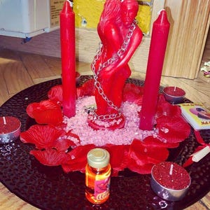 Puede incluir: Una composici&oacute;n rom&aacute;ntica con velas rojas, una estatua roja de figuras abrazadas y p&eacute;talos de rosa rojos sobre un plato negro. Tambi&eacute;n hay peque&ntilde;as velas y una botella de l&iacute;quido.