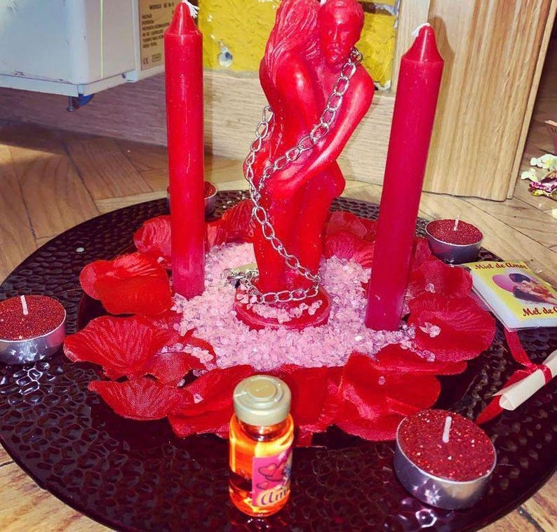 Puede incluir: Una composici&oacute;n rom&aacute;ntica con velas rojas, una estatua roja de figuras abrazadas y p&eacute;talos de rosa rojos sobre un plato negro. Tambi&eacute;n hay peque&ntilde;as velas y una botella de l&iacute;quido.