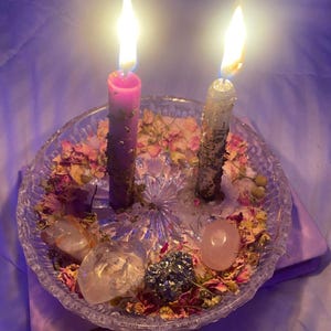 Puede incluir: Dos velas encendidas, una rosa y otra gris, en un plato de cristal transparente. El plato contiene flores secas, cristales y una tela rosa. Las velas arden brillantemente.