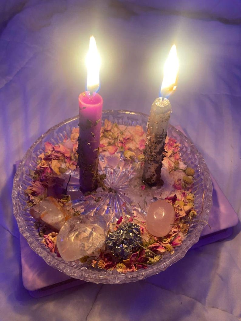 Puede incluir: Dos velas encendidas, una rosa y otra gris, en un plato de cristal transparente. El plato contiene flores secas, cristales y una tela rosa. Las velas arden brillantemente.
