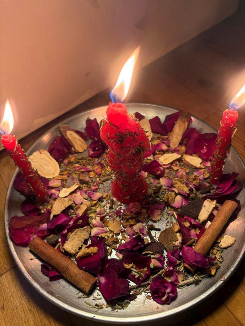 Puede incluir: Un plato plateado contiene tres velas rojas encendidas, rodeadas de p&eacute;talos de rosa secos, hierbas y palitos de canela. La vela central tiene una forma texturizada &uacute;nica. Las llamas crean un ambiente m&iacute;stico.