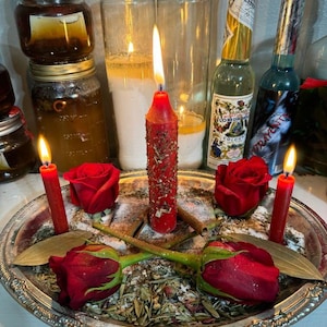 Puede incluir: Una bandeja plateada contiene rosas rojas, velas y hierbas. Dos peque&ntilde;as velas rojas flanquean una vela roja central texturizada. Dos botellas de vidrio y varios frascos est&aacute;n en el fondo. La escena est&aacute; iluminada por las llamas de las velas.