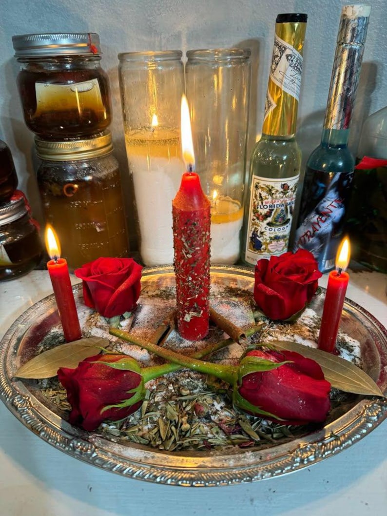 Puede incluir: Una bandeja plateada contiene rosas rojas, velas y hierbas. Dos peque&ntilde;as velas rojas flanquean una vela roja central texturizada. Dos botellas de vidrio y varios frascos est&aacute;n en el fondo. La escena est&aacute; iluminada por las llamas de las velas.
