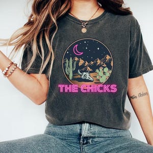 Op de afbeelding: Donkergrijs T-shirt met een woestijnontwerp. Het ontwerp toont een halve maan, sterren, bergen, cactussen en een schedel, met de tekst "THE CHICKS" in roze neonletters. Het shirt wordt gecombineerd met een blauwe jeans en witte schoenen.