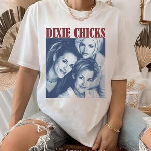 Pode incluir: T-shirt branco com o texto bordô "DIXIE CHICKS" acima de uma imagem em tons de azul de três mulheres. A t-shirt é de estilo casual e unissexo.