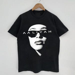 May include: Black t-shirt featuring a black and white image of a person wearing sunglasses. The word "AALIYAH" is printed in white above the sunglasses. The t-shirt is hanging on a wooden hanger.