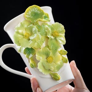 Könnte beinhalten: Weiße Keramiktasse mit Henkel, verziert mit 3D-Blumenverzierungen in Grün und Gelb. Die Blumen haben detaillierte Blütenblätter und gelbe Zentren. Die Tasse wird vor einem schwarzen Hintergrund gehalten.
