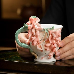 Könnte beinhalten: Eine weiße Keramik-Teetasse mit detailliertem Blumendesign. Die Tasse hat einen hellgrünen Henkel und ist mit rosa- und pfirsichfarbenen Blumen verziert. Die Tasse wird von zwei Händen gehalten.