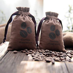 Op de afbeelding: Twee bruine zakjes met trekkoord met afbeeldingen van koffiebonen en de woorden "Coffee fragrance" liggen op een houten oppervlak bezaaid met koffiebonen. De zakken hebben donkerpaarse linten.