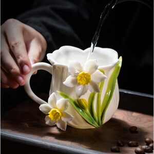 Könnte beinhalten: Eine weiße Keramiktasse mit Henkel, verziert mit 3D-Osterglockenblumen. Die Blumen haben weiße Blütenblätter und gelbe Zentren, mit grünen Blättern. Wasser wird in die Tasse gegossen. Die Tasse steht auf einem Holztablett.