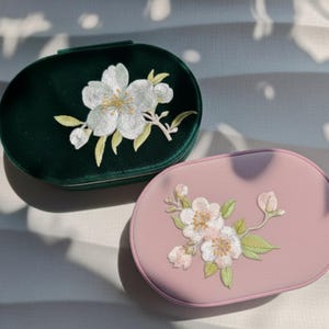 May include: Two oval-shaped cosmetic cases. One is dark green velvet with white floral embroidery. The other is light pink with similar white floral embroidery. Both cases have a zipper closure.