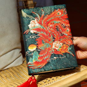 May include: A teal-colored journal with an embroidered phoenix design in red, orange, and teal. The phoenix is surrounded by white and orange flowers, with a yellow moon and cloud details. Red tassels hang from the bottom.