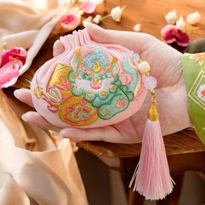 Puede incluir: Una bolsa rosa bordada con un diseño detallado que presenta una criatura mítica en tonos de azul, verde y dorado. La bolsa tiene una borla rosa y un dije de cuentas. El fondo incluye flores y una superficie de madera.