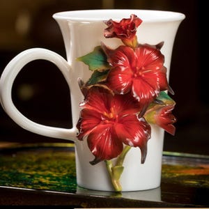 Könnte beinhalten: Weißer Keramikbecher mit gebogenem Henkel, verziert mit dreidimensionalen roten Hibiskusblüten und grünen Blättern. Die Blüten haben ein glänzendes Finish und verleihen dem floralen Design Eleganz.