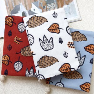 May include: Three folded fabric pieces in red, white, and blue, each adorned with embroidered leaf patterns in shades of brown, orange, and white. The leaves vary in size and design, creating a natural, autumnal theme.