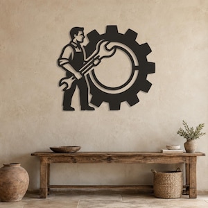 May include: A black metal wall art piece featuring a silhouette of a mechanic wearing an apron and holding a large wrench, positioned in front of a large gear. The design is flat, monochromatic, and depicts a worker standing beside a cog.