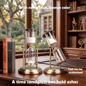 May include: Two bronze-colored metal hourglasses with clear glass chambers containing white sand. One hourglass stands upright, while the other is tilted on a swing base. The text reads: With swing base, bronze color, Text can be customized, A time sandglass can hold ashes.