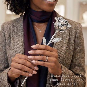May include: A polished silver-colored stainless steel rose flower with a long stem and detailed leaves. The metal rose features a hollow design intended to hold ashes. The text on the image reads: Stainless steel rose flower, can hold ashes.