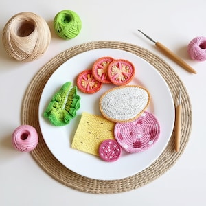 Könnte beinhalten: Ein weißer Teller mit gehäkelten Lebensmitteln, darunter Tomatenscheiben, Salat, ein Spiegelei, Käse und eine Salamischeibe. Der Teller steht auf einem gewebten Platzset, mit Garn und Häkelnadeln in der Nähe. Die Lebensmittel sind in verschiedenen Farben.