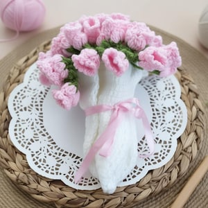May include: A handmade bouquet of pink crochet flowers with green stems, wrapped in white crochet and tied with a pink ribbon. The bouquet rests on a white doily and a woven placemat, with a ball of pink yarn in the background.