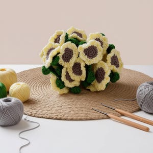 May include: A handmade crochet sunflower bouquet with yellow petals and brown centers. The bouquet sits on a woven mat, surrounded by balls of yarn in yellow, green, and gray. Two crochet hooks are also visible.