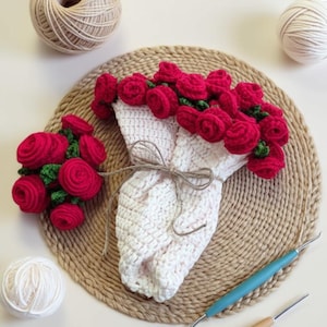 May include: A crocheted bouquet of red roses wrapped in white yarn, tied with twine. A smaller bouquet of red roses is to the left. The bouquets rest on a woven, circular placemat. Two balls of yarn and two crochet tools are also visible.