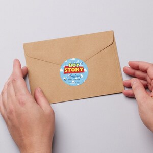 May include: A brown envelope with a round sticker that reads "It's a Boy Story" in blue, red, and white. The sticker is for Sebastian's baby shower. The envelope is held by two hands.