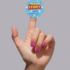 May include: A round baby shower sticker with the text "It's a Boy Story" in blue, red, and yellow. The sticker includes the name "Jacob's" and the date "October 23, 20xx" on a light blue background with clouds.