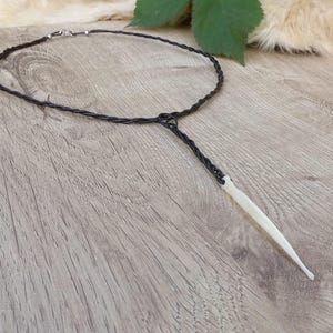 May include: A black cord necklace with a pendant. The necklace features a braided cord design and a white, elongated pendant. The necklace is displayed on a wooden surface.