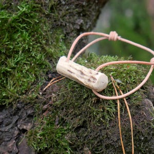May include: A handmade antler pendant with a light brown leather cord. The pendant is a light tan color with darker brown markings. The cord is knotted at the top and is resting on a bed of green moss.