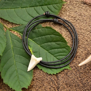 Op de afbeelding: Een zwarte leren koordketting met een crèmekleurige tandhanger ligt op groene bladeren. De ketting is bovenaan geknoopt en heeft een eenvoudig, natuurlijk ontwerp. De achtergrond is een bruin, getextureerd oppervlak.
